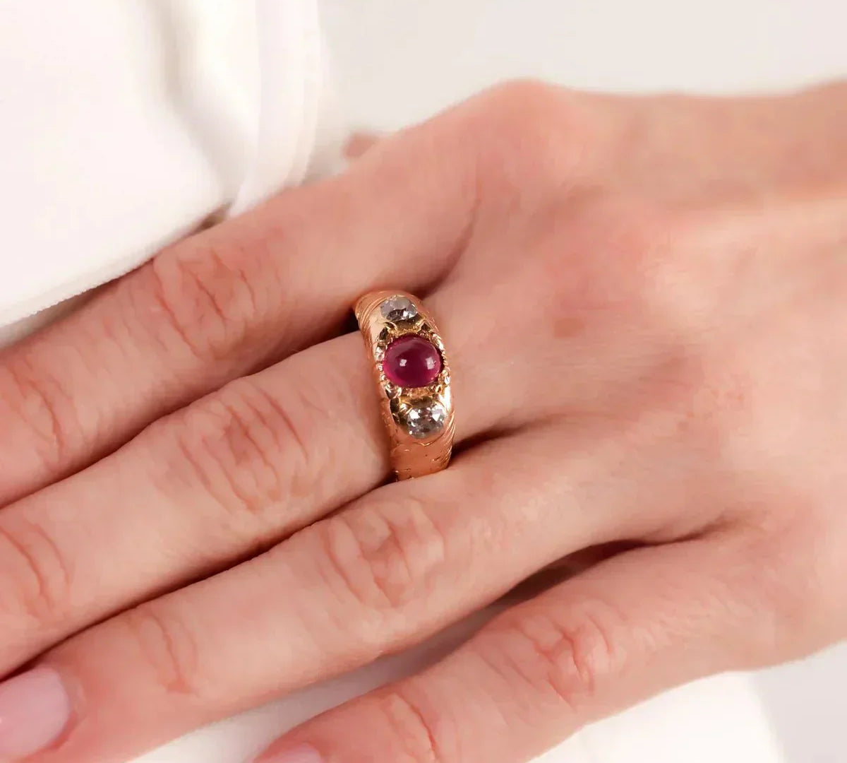 Victorian-era gold ring featuring a cabochon ruby flanked by two rose-cut diamonds, shown on a hand for scale. The antique design highlights intricate metalwork and timeless elegance