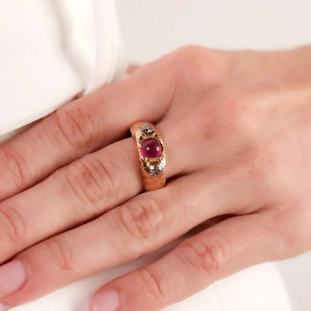 Victorian-era gold ring featuring a cabochon ruby flanked by two rose-cut diamonds, shown on a hand for scale. The antique design highlights intricate metalwork and timeless elegance