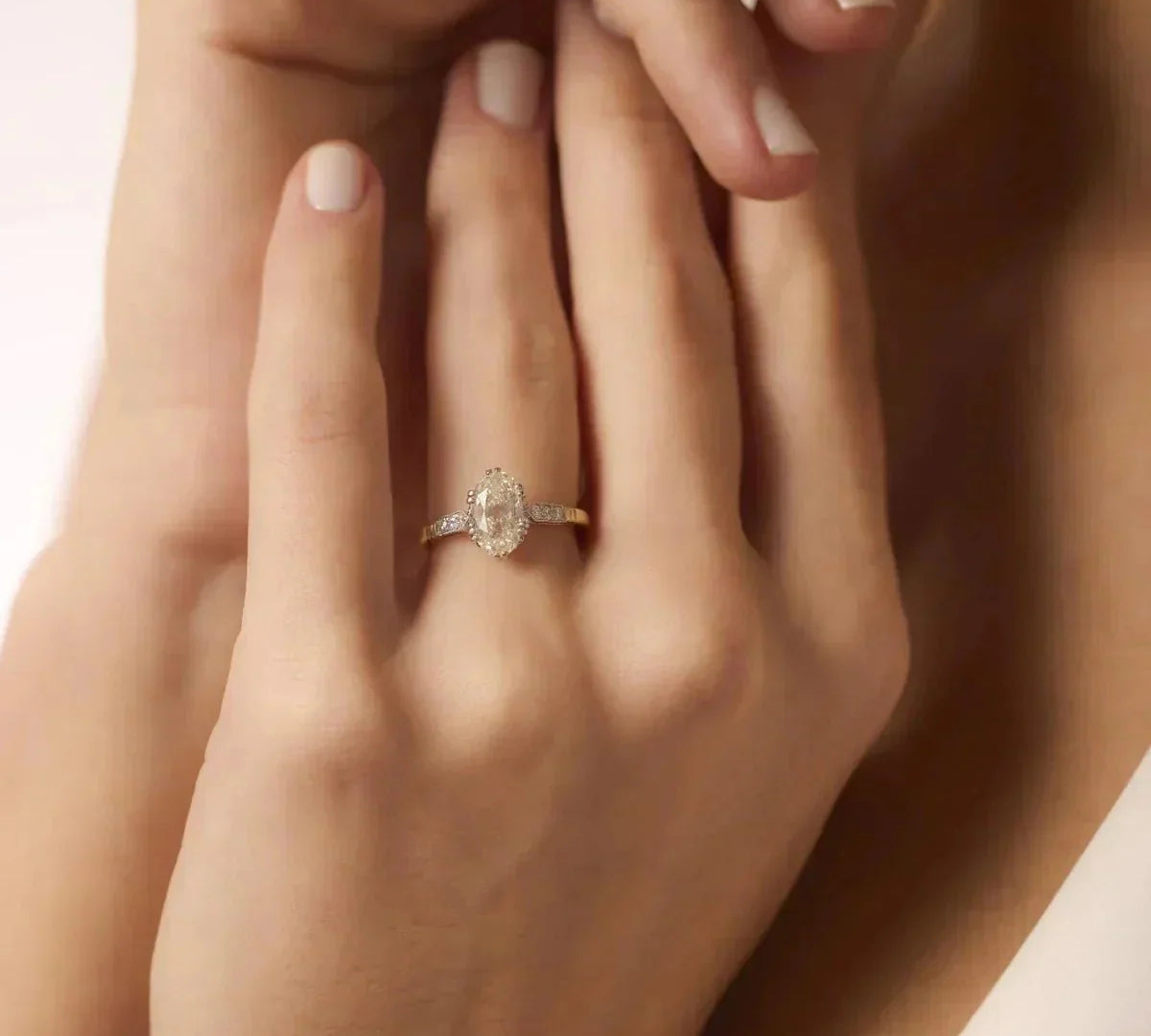 Elegant oval-shaped diamond engagement ring set in yellow gold with diamond-accented shoulders, displayed on a woman's hand with a soft, romantic pose