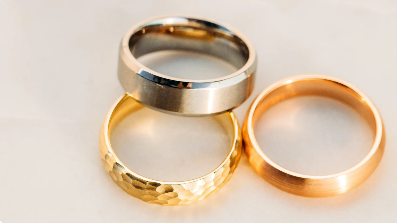 Three men’s wedding bands in white, yellow, and rose gold finishes, displayed together as examples of styles in Trumpet & Horn’s Wedding Ring Buying Guide.