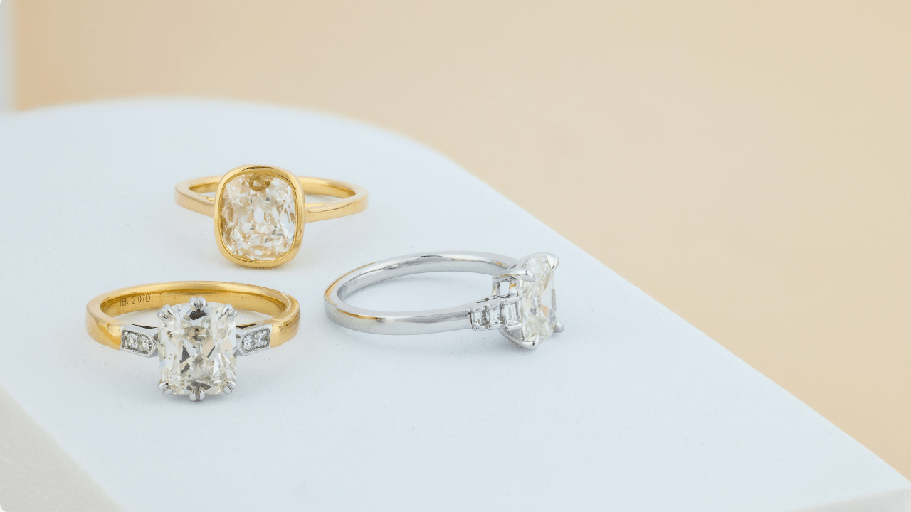Three vintage engagement rings displayed on a white surface, featuring yellow gold and platinum settings with cushion and emerald-cut diamonds.