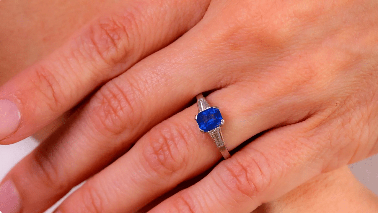Close-up of a hand wearing a vintage platinum ring featuring a vivid blue sapphire flanked by two tapered baguette diamonds.